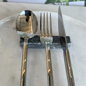 Può includere: Un set di posate in acciaio inossidabile, composto da un cucchiaio, una forchetta e un coltello. Le posate sono appoggiate su un poggia posate in marmo grigio scuro. Il coltello ha una lama dritta. Il set è su una superficie di marmo bianco.