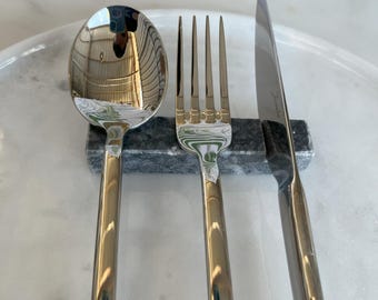 May include: A stainless steel spoon, fork, and knife set. The silverware is resting on a dark gray marble utensil rest. The spoon and fork have four tines. The knife has a straight blade. The set is on a white marble surface.