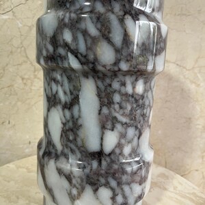 May include: A cylindrical marble vase with a polished finish. The vase features a pattern of white and dark gray veining. The design includes a series of horizontal bands, creating a textured appearance. The vase is suitable for home decor.