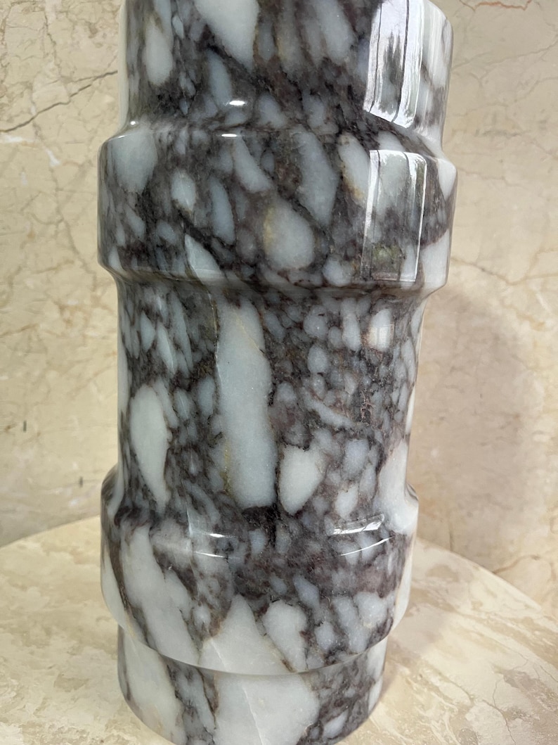 May include: A cylindrical marble vase with a polished finish. The vase features a pattern of white and dark gray veining. The design includes a series of horizontal bands, creating a textured appearance. The vase is suitable for home decor.