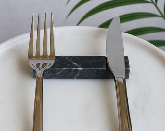 May include: A gold-colored fork and a silver knife rest on a white marble plate. A black marble knife rest holds the knife. The fork and knife are part of a dining set. Green leaves are in the background.