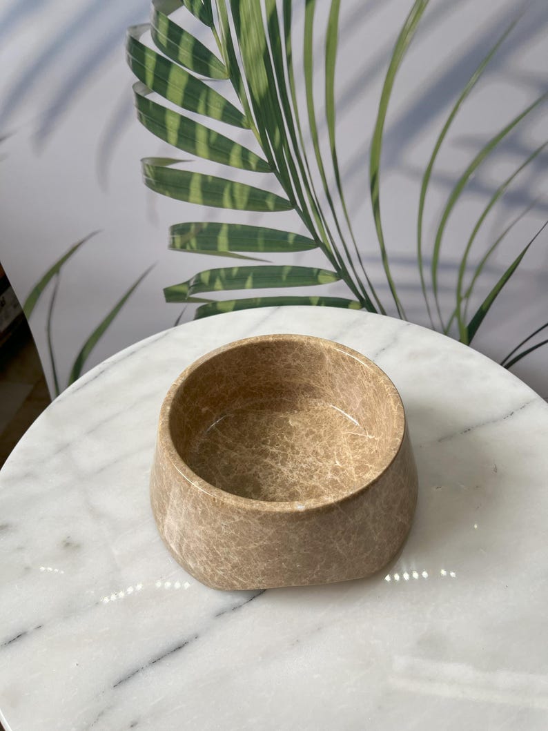 Puede incluir: Un cuenco peque&ntilde;o y redondo de color beige con una superficie texturizada. El cuenco tiene una abertura ancha y una base ligeramente c&oacute;nica. El cuenco est&aacute; sobre una mesa de m&aacute;rmol blanco. El fondo presenta un patr&oacute;n de hojas verdes y blancas.