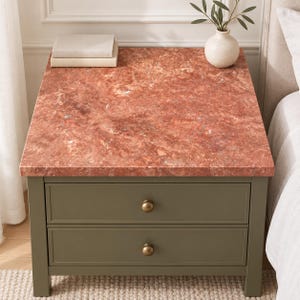 May include: A square bedside table with a reddish-brown marble top and an olive green wooden base featuring two drawers with round brass-coloured knobs. Two stacked beige books and a small white vase with green leaves sit on the surface.