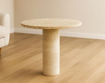 May include: A round, beige side table with a cylindrical base. The table is made of a light-colored stone material and stands approximately 20 inches tall. The table is a neutral color and would fit in many home decor styles.