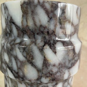 May include: A cylindrical marble vase with a unique shape. The vase features a white and dark gray marbled pattern. The design includes a distinct, slightly indented band around the middle, adding a touch of elegance to the overall aesthetic.
