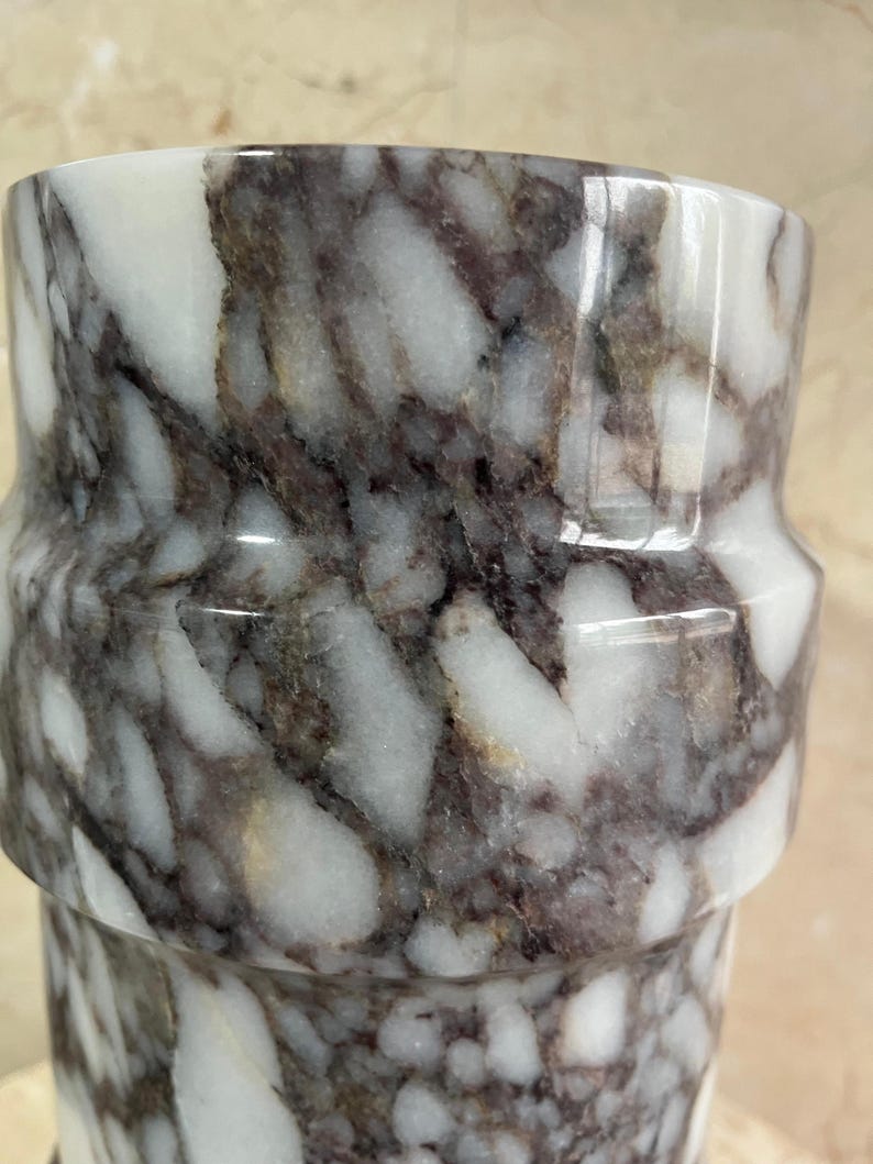 May include: A cylindrical marble vase with a unique shape. The vase features a white and dark gray marbled pattern. The design includes a distinct, slightly indented band around the middle, adding a touch of elegance to the overall aesthetic.