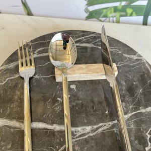 May include: A stainless steel fork, spoon, and knife set arranged on a dark gray marble-patterned surface. The silverware has a polished, reflective finish. The spoon rests on a small, light-colored rectangular block. The background features a white surface and green plant.