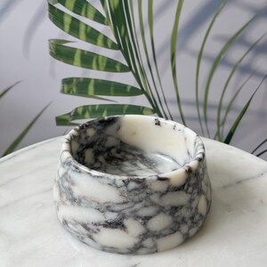 Puede incluir: Un peque&ntilde;o cuenco redondo de m&aacute;rmol con un patr&oacute;n veteado blanco y gris. El cuenco est&aacute; sobre una superficie de m&aacute;rmol blanco. Hojas de palmera verdes y blancas en el fondo.