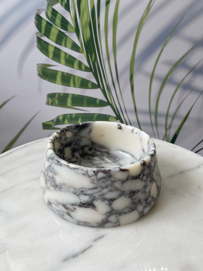 Puede incluir: Un peque&ntilde;o cuenco redondo de m&aacute;rmol con un patr&oacute;n veteado blanco y gris. El cuenco est&aacute; sobre una superficie de m&aacute;rmol blanco. Hojas de palmera verdes y blancas en el fondo.