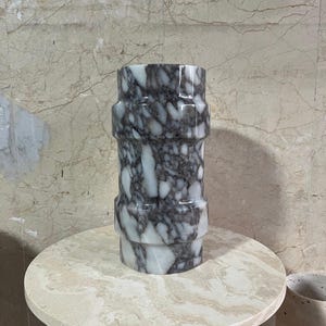 May include: A cylindrical marble vase with a gray and white pattern. The vase has a series of horizontal bands and sits on a round, light-colored table. The background features a marble wall.