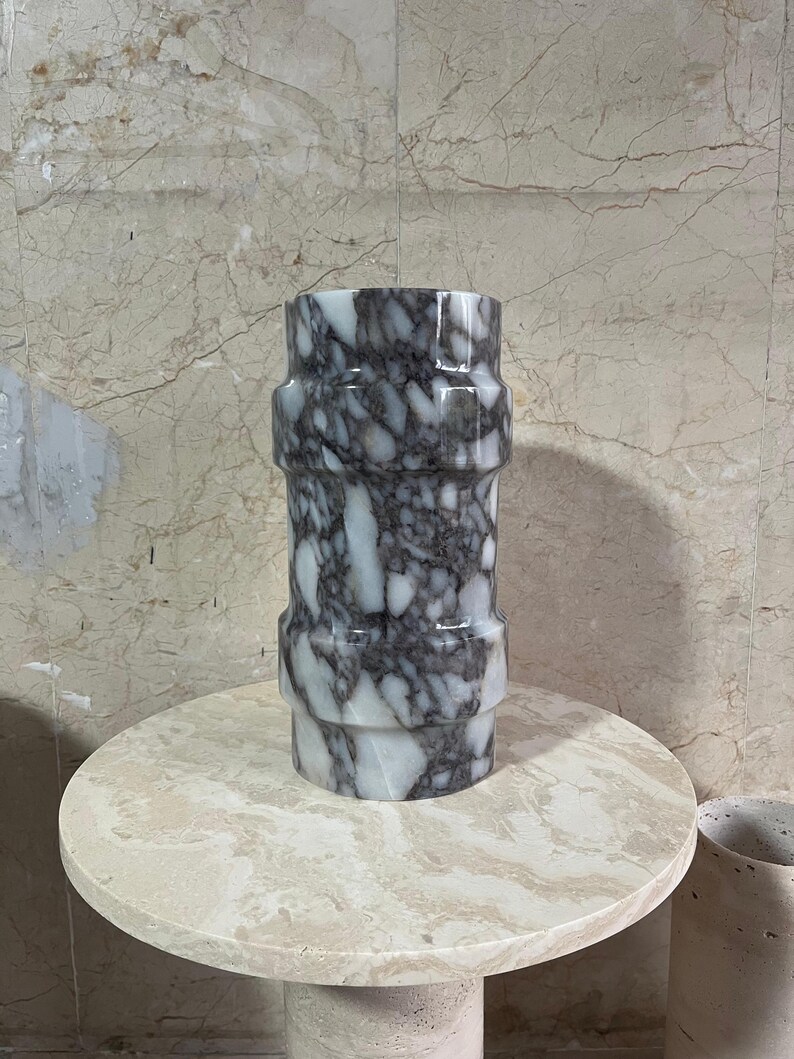 May include: A cylindrical marble vase with a gray and white pattern. The vase has a series of horizontal bands and sits on a round, light-colored table. The background features a marble wall.