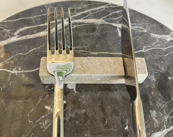 May include: A stainless steel fork and knife set resting on a gray stone holder. The fork has four tines and a gold-colored handle. The knife has a long, slender blade and a matching handle. The set is placed on a dark gray marble surface.