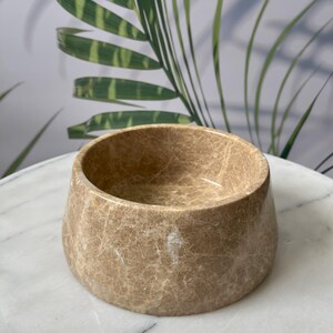 Puede incluir: Un cuenco redondo beige con una superficie texturizada. El cuenco tiene una abertura ancha y una base ligeramente c&oacute;nica. El cuenco est&aacute; hecho de un material similar a la piedra y se encuentra sobre una superficie blanca.