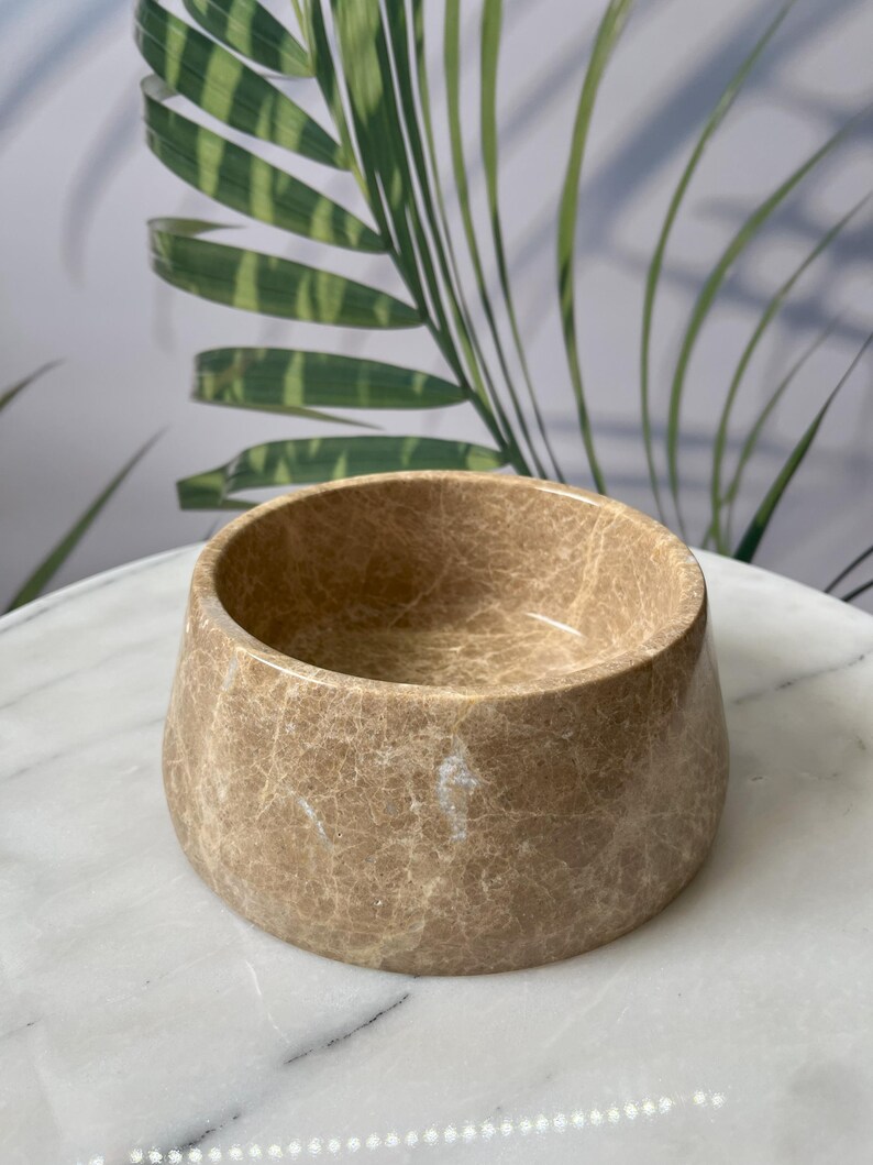 Puede incluir: Un cuenco redondo beige con una superficie texturizada. El cuenco tiene una abertura ancha y una base ligeramente c&oacute;nica. El cuenco est&aacute; hecho de un material similar a la piedra y se encuentra sobre una superficie blanca.