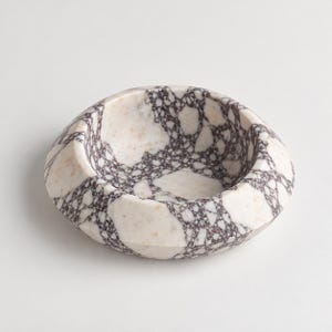 May include: A circular stone bowl featuring a polished surface with a distinct brecciated pattern. The design consists of large, irregular white and cream-colored stone fragments interconnected by a dark, intricate web of deep purple veining.