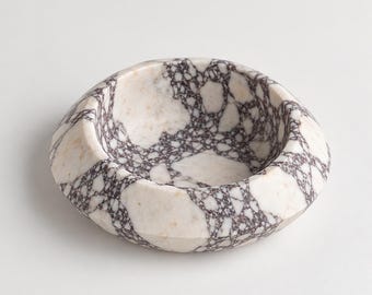 Puede incluir: Un cuenco circular de piedra con una superficie pulida y un patrón brechoide distintivo. El diseño consta de grandes fragmentos de piedra irregulares de color blanco y crema, interconectados por una red oscura y compleja de vetas moradas.