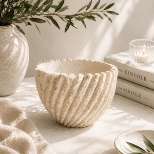 May include: A beige stone bowl with a textured, spiraling ribbed exterior and a wide, open circular rim. The bowl features a rough, porous surface finish and a deep, rounded interior basin, presented as a standalone decorative vessel.