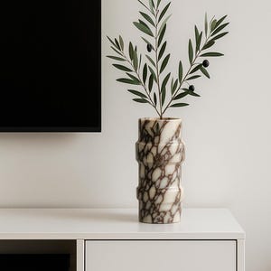 May include: A decorative marble vase with a brown and white pattern, holding a sprig of olive branches with green leaves and black olives. The vase has a cylindrical shape with horizontal ridges. Home decor.