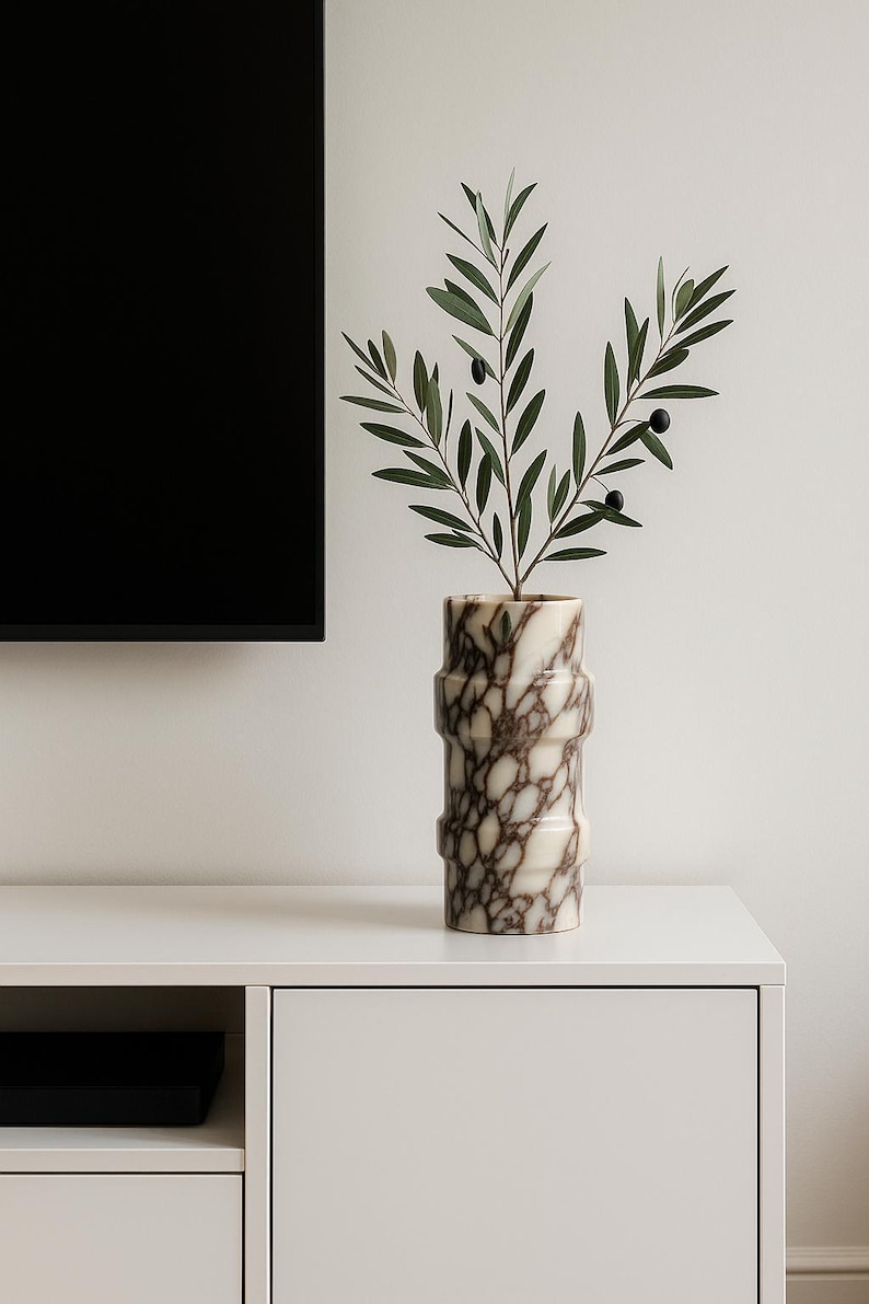 May include: A decorative marble vase with a brown and white pattern, holding a sprig of olive branches with green leaves and black olives. The vase has a cylindrical shape with horizontal ridges. Home decor.