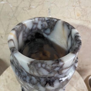 May include: A marble vase with a swirling pattern of white, gray, and brown. The vase has a wide, rounded top and a slightly flared base. The polished surface reflects light, highlighting the natural veining of the stone. A decorative item for the home.