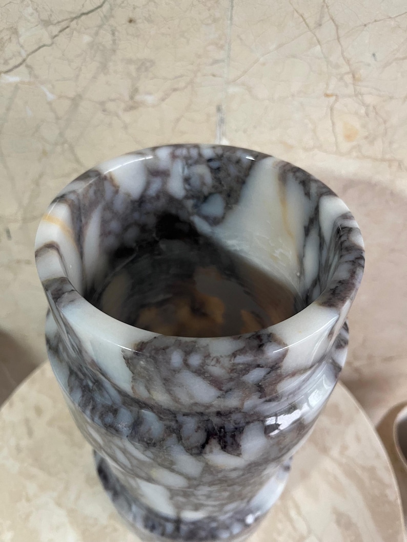 May include: A marble vase with a swirling pattern of white, gray, and brown. The vase has a wide, rounded top and a slightly flared base. The polished surface reflects light, highlighting the natural veining of the stone. A decorative item for the home.