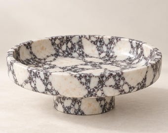 May include: A circular pedestal bowl featuring a distinctive brecciated pattern with large, irregular white and cream-colored stone fragments embedded within a dark, charcoal-colored matrix. The bowl has a shallow basin and a cylindrical base.