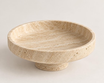 May include: A circular beige travertine stone bowl with a porous, textured surface and natural veining patterns. The bowl features a shallow basin and is supported by a thick, cylindrical pedestal base, creating a raised, elevated presentation.