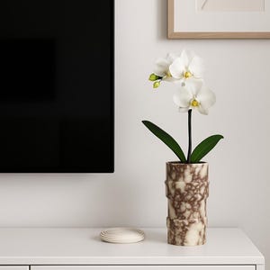 May include: A white orchid plant with green leaves in a decorative brown and cream-colored vase. The vase has a textured, cylindrical design. A stack of white coasters sits to the left of the vase. The scene is set on a white cabinet.