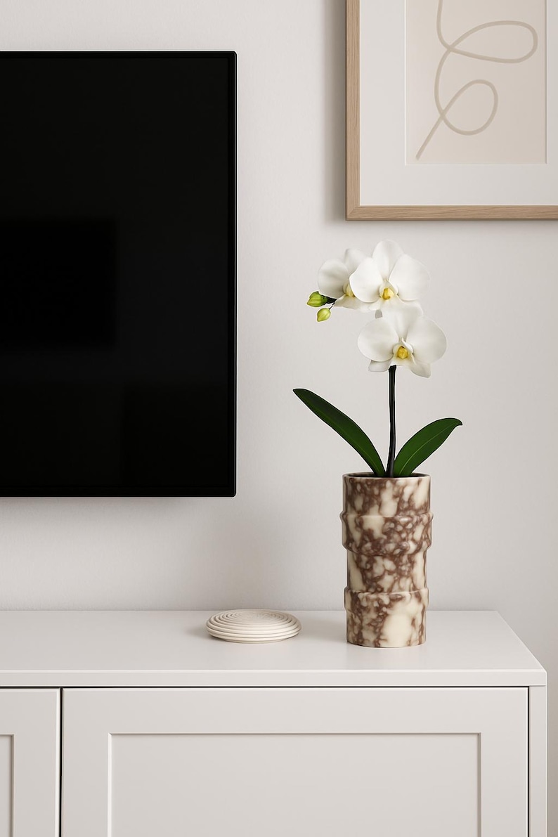 May include: A white orchid plant with green leaves in a decorative brown and cream-colored vase. The vase has a textured, cylindrical design. A stack of white coasters sits to the left of the vase. The scene is set on a white cabinet.