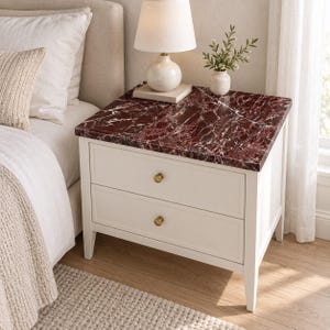 Peut inclure: Une table de chevet en bois blanc dotée de deux tiroirs avec de petits boutons ronds en laiton et un plateau rectangulaire au motif de marbre rouge foncé et blanc. Le meuble repose sur quatre pieds et est placé à côté d'un lit.