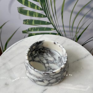Puede incluir: Un peque&ntilde;o cuenco redondo de m&aacute;rmol con un patr&oacute;n blanco y gris. El cuenco est&aacute; sobre una superficie de m&aacute;rmol blanco, con hojas de palmera verdes en el fondo. El cuenco tiene aproximadamente 7,6 cm de di&aacute;metro y 5 cm de alto.