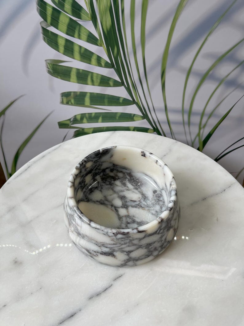 Puede incluir: Un peque&ntilde;o cuenco redondo de m&aacute;rmol con un patr&oacute;n blanco y gris. El cuenco est&aacute; sobre una superficie de m&aacute;rmol blanco, con hojas de palmera verdes en el fondo. El cuenco tiene aproximadamente 7,6 cm de di&aacute;metro y 5 cm de alto.