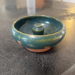 Può includere: Una piccola ciotola in ceramica color verde acqua con un centro rialzato. La ciotola ha una finitura rustica leggermente irregolare e un bordo marrone.