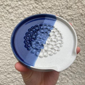 May include: A round ceramic spoon rest featuring a two-tone design with a glossy deep blue glaze on one side and a speckled off-white glaze on the other. The centre displays a raised, textured floral pattern that spans across both colour sections.
