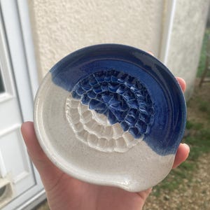 May include: A handmade ceramic spoon rest. The spoon rest is half white and half blue, with a deep blue textured centre. The spoon rest is held in a hand. The spoon rest is approximately 15 cm in diameter.