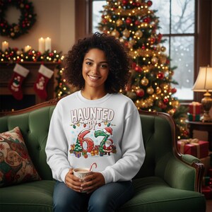 May include: A white sweatshirt with the text "HAUNTED BY 67" in red and green, with Christmas lights and Santa Claus graphics. The person is sitting on a green couch in front of a Christmas tree.