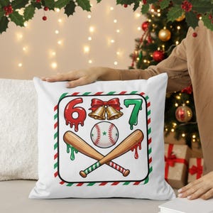 May include: White square pillow with a baseball-themed Christmas design. The design includes the numbers 6 and 7, a baseball, crossed bats, and bells. The numbers and dripping accents are red and green.