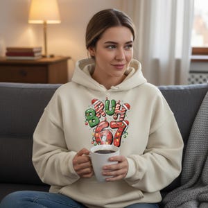 May include: A cream-colored hoodie with the text "Bruh 67" in a festive, Christmas-themed design. The text features green and red colors with candy cane accents and Christmas lights. The person is holding a white mug.