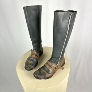 Puede incluir: Un par de botas negras de cuero de caña alta con detalles en cuero marrón y una cremallera lateral. Las botas tienen un tacón grueso y una correa alrededor del tobillo.