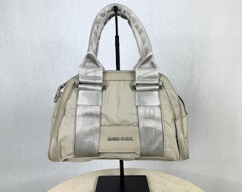 May include: A beige Diesel handbag with silver accents. The bag features a structured design with a top handle and silver straps. The word "DIESEL" is visible on the front of the bag.