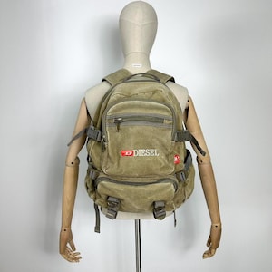 Può includere: Uno zaino Diesel color cachi con più scomparti e spallacci regolabili. Lo zaino presenta il logo Diesel in rosso e bianco sul davanti. La borsa è esposta su un manichino.