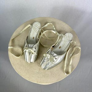 May include: A pair of silver ballet flats with cream-colored ribbon ties. The shoes have a pointed toe and a small bow detail. The insoles are white with a quilted pattern. The shoes are displayed on a round, beige surface.