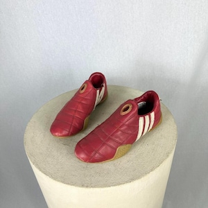 May include: A pair of red athletic shoes with white stripes and a gold-colored accent on the top. The shoes are made of a quilted material and have a tan sole. The shoes are displayed on a white pedestal.