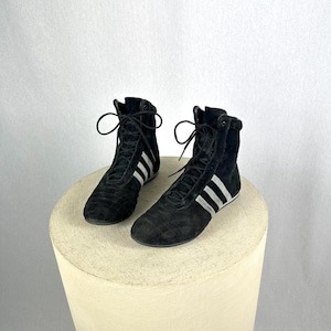 Puede incluir: Un par de zapatillas altas de ante negro con rayas blancas. Las zapatillas tienen cordones y se exhiben sobre un pedestal beige. Las zapatillas tienen un diseño deportivo clásico.