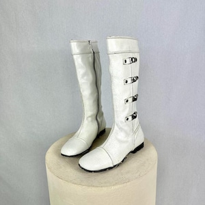 Puede incluir: Un par de botas blancas hasta la rodilla con suelas negras. Una bota tiene una cremallera, la otra tiene tres hebillas. Las botas están hechas de un material texturizado y se exhiben sobre un pequeño pedestal beige redondo.