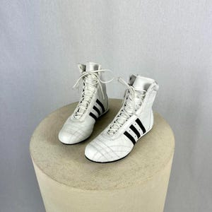 Puede incluir: Un par de zapatillas altas blancas con rayas negras en los laterales. Las zapatillas tienen un diseño con cordones y están hechas de un material liso. Los zapatos se exhiben sobre un pedestal redondo de color neutro.