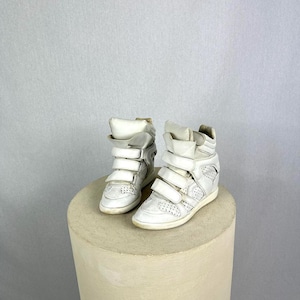 May include: A pair of white high-top sneakers with multiple hook-and-loop straps. The sneakers have a wedge heel and are made of a textured material. The shoes are displayed on a cylindrical, light-colored pedestal.