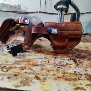 Puede incluir: Una funda de cuero marrón con forma moldeada, un poste de metal plateado y una correa de nailon negro con hebilla de plástico. La funda está construida con costuras visibles y un cierre de botón a presión de metal en la correa lateral.