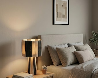 Puede incluir: Lámpara de mesa con pantalla cilíndrica a rayas verticales en tonos negros y naturales. La lámpara está sobre una mesa de madera junto a una cama con almohadas y una obra de arte enmarcada. La pantalla mide unos 30 cm de alto.