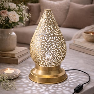 Op de afbeelding: Een goudkleurige, druppelvormige tafellamp met ingewikkelde uitgesneden patronen. De lamp heeft een ronde voet en een aan/uit-schakelaar. De lamp is ongeveer 30 cm hoog en werpt een decoratief lichtpatroon.
